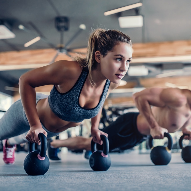 Femme et homme faisant des pompes sur kettlebells dans une salle de sport, regard concentré.