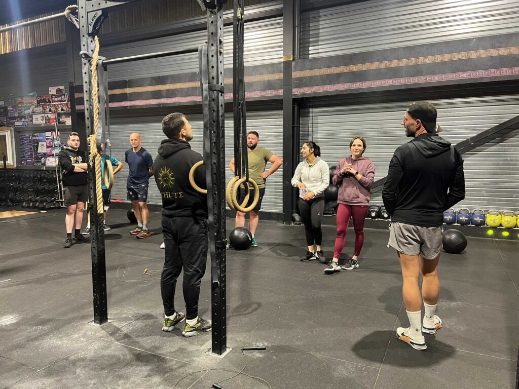 Groupe d'athlètes écoutant un coach dans une salle de CrossFit avec rig et rings. Texte visible : HLTΣ.