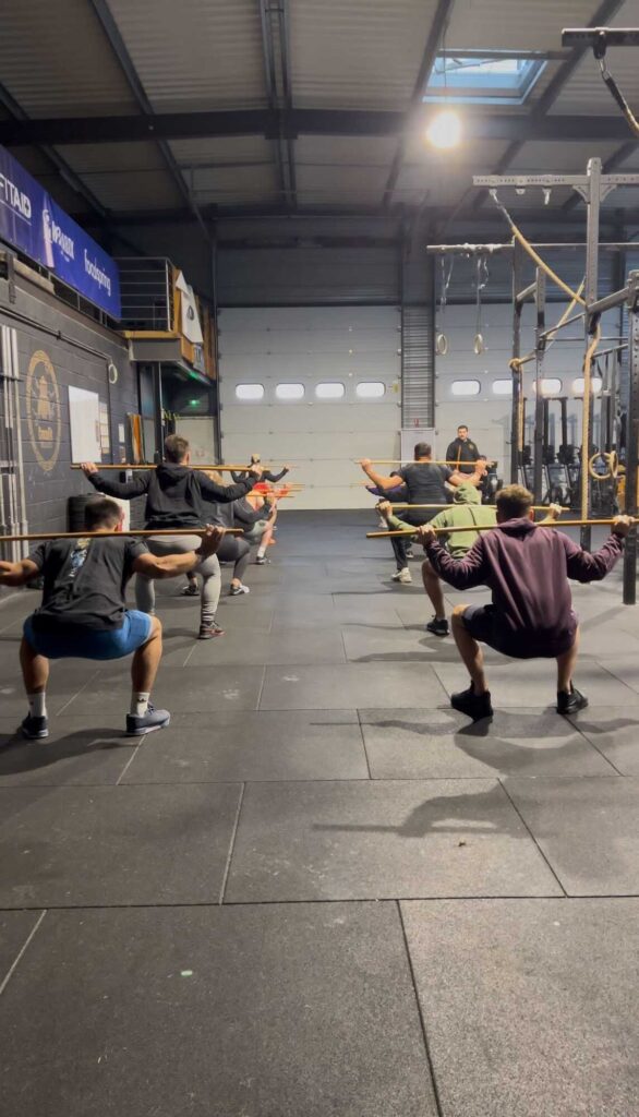 Squats avec bâtons en groupe dans un gym type CrossFit. Instructeur en arrière plan. Logos FITAD, FOODSPRING visibles.