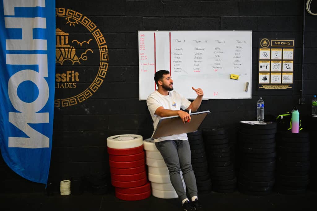 Coach CrossFit Iliades explique l'entraînement d'équipe. Tableau des scores, règles de la Box et poids visibles sur fond noir.